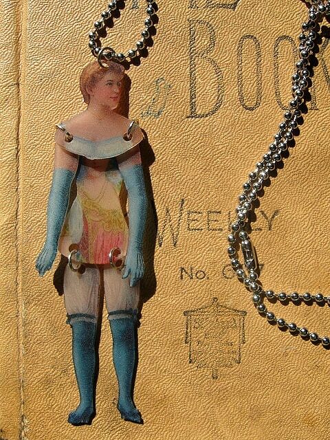 Art Doll Necklace in Blue. Created from shrink plastic and coated to protect it. Torso was hand cut from a scanned vintage label that was printed on shrink plastic as well. Arms and legs are jointed so she moves and dances as you wear her.